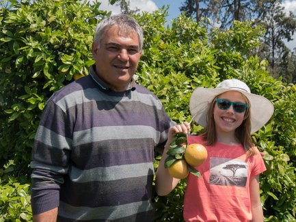 Ein Mann und ein Mädchen stehen vor einem grünen Strauch, während das Mädchen stolz zwei Orangen in der Hand hält.
