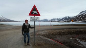 Ein Mann steht neben einem Warnschild, das vor Eisbären warnt, mit einer beeindruckenden Berglandschaft im Hintergrund.