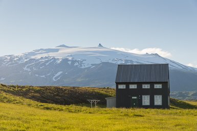 Ein schwarzes Sommerhaus steht in einer grünen Wiese, mit schneebedeckten Bergen im Hintergrund, die unter blauem Himmel leuchten.