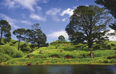 Eine malerische Landschaft mit sanften Hügeln, bunten Hobbit-Höhlen und einem ruhigen Gewässer im Vordergrund.
