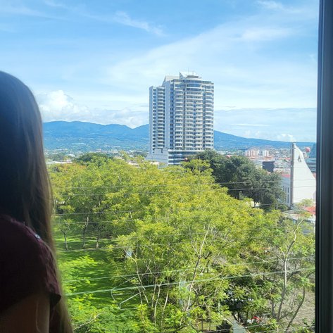 Eine Person mit langen Haaren blickt aus einem Fenster auf eine grüne Landschaft und moderne Gebäude in der Ferne.