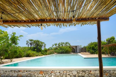 Ein klarer Pool spiegelt den blauen Himmel wider, umgeben von üppigem Grün und einem rustikalen Holzüberstand.
