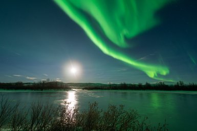 Ein atemberaubendes Nordlicht erhellt den Nachthimmel über einem gefrorenen See, während der Mond sanft reflektiert.