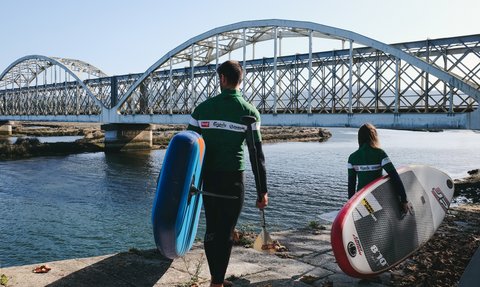 Ein Mann und ein Kind stehen am Wasser, bereit zum Paddeln, während eine Brücke im Hintergrund sichtbar ist.