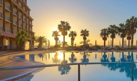 Ein atemberaubender Sonnenuntergang spiegelt sich im glitzernden Pool eines Hotels, umgeben von Palmen und Liegen.