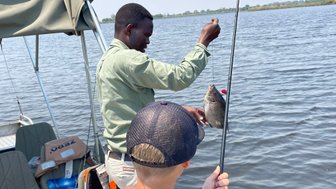 Ein Angler hält stolz einen gefangenen Fisch über dem Wasser, während ein Junge gespannt zuschaut.