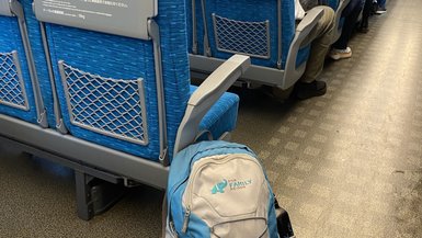 Ein blauer Rucksack steht auf dem Boden eines Zugabteils, umgeben von blauen Sitzen und Passagieren im Hintergrund.