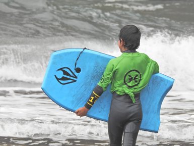 Ein Junge in einem grünen Neoprenanzug trägt ein blaues Surfbrett und blickt auf die Wellen des Ozeans.