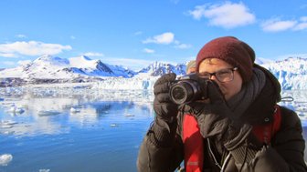 Eine Person mit einer Kamera steht am Wasser und fotografiert die beeindruckende Berglandschaft im Hintergrund.