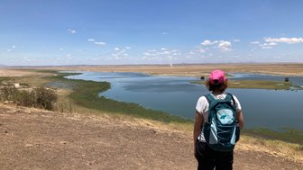 Eine Person mit einem Rucksack steht am Ufer eines ruhigen Gewässers und blickt auf die weite Landschaft von Amboseli.