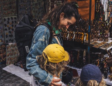 Eine lächelnde Frau mit langen, lockigen Haaren umarmt zwei Kinder, während sie an einem bunten Markt stehen.