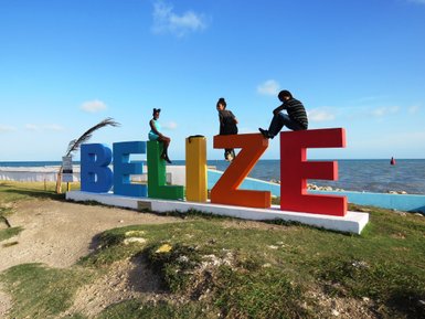 Drei Personen sitzen auf bunten Buchstaben, die das Wort 'BELIZE' bilden, vor einem klaren blauen Himmel und dem Meer.