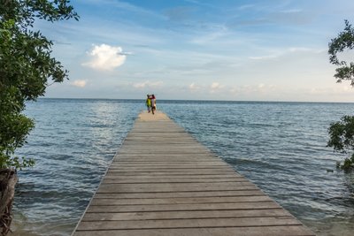 Zwei Personen gehen auf einem Holzsteg, umgeben von Wasser und Bäumen, unter einem klaren Himmel mit wenigen Wolken.