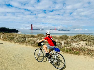 Ein Junge in einem roten T-Shirt fährt mit dem Fahrrad entlang eines Küstenwegs, mit der Golden Gate Bridge im Hintergrund.