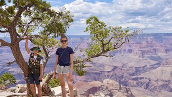 Ein Junge mit Sonnenbrille und Hut steht an einem Baum, während eine Frau in sportlicher Kleidung lächelt, mit dem Grand Canyon im Hintergrund.