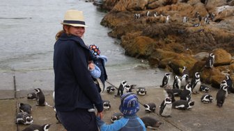 Eine Frau mit einem Hut hält ein Baby, während ein Junge an ihrer Hand steht und Pinguine im Hintergrund beobachtet.