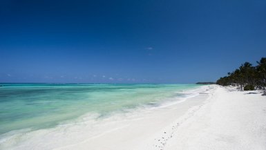 Ein endloser, weißer Sandstrand erstreckt sich unter einem strahlend blauen Himmel, während sanfte Wellen das Ufer umspülen.