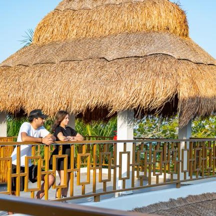 Ein Paar lehnt an einem Holzgeländer und genießt die Aussicht auf das Wasser und die umgebende Natur unter einem Strohdach.