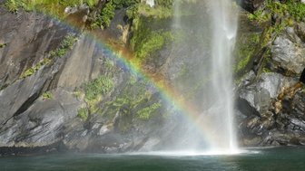 Ein Wasserfall stürzt über eine felsige Klippe, während ein Regenbogen in der Gischt leuchtet und die grüne Vegetation umrahmt.