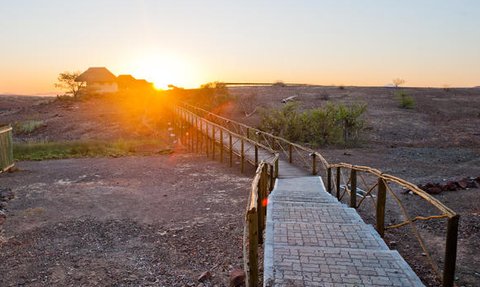 Die Sonne geht hinter einer hügeligen Landschaft unter, während ein Holzsteg zur Unterkunft führt.
