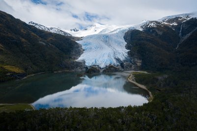 Ein majestätischer Gletscher fließt in einen ruhigen See, umgeben von bewaldeten Bergen und schneebedeckten Gipfeln.