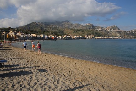 Ein sonniger Strand in Giardini Naxos, wo Menschen am Wasser spazieren und die sanften Wellen genießen.