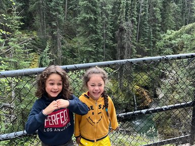 Zwei fröhliche Kinder stehen an einem Geländer, umgeben von hohen, grünen Bäumen und einem klaren Fluss im Hintergrund.