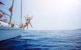 Drei fröhliche Personen springen von einem Segelboot ins glitzernde blaue Wasser, umgeben von strahlendem Sonnenschein.