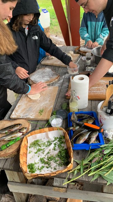 Eine Gruppe von Kindern und Erwachsenen bereitet frischen Fisch und Teig auf einem Holztisch im Freien zu.