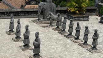 Eine Gruppe von zehn steinernen Figuren steht auf einem Platz, umgeben von üppigem Grün und historischen Gebäuden im Hintergrund.