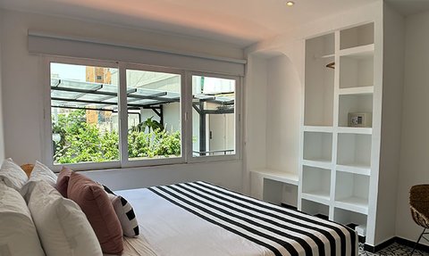 Ein helles Schlafzimmer mit einem großen Fenster, das auf einen grünen Innenhof blickt, und einem Bett mit gestreifter Bettwäsche.