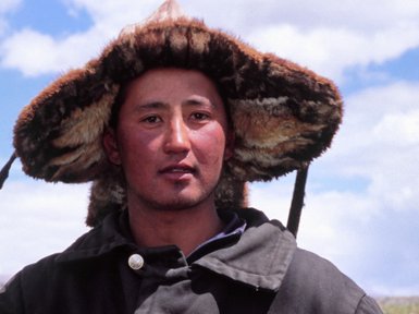 Ein mongolischer Mann trägt einen traditionellen Hut und sitzt mit zwei Kindern in einer grünen Landschaft unter einem blauen Himmel.
