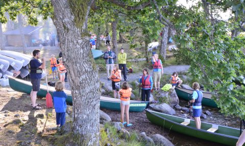 Eine Gruppe von Menschen in Schwimmwesten bereitet sich auf ein Kanuabenteuer am Ufer eines ruhigen Sees vor.