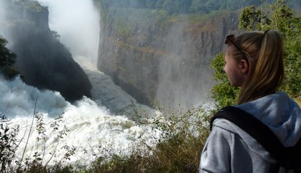 Eine junge Frau steht am Rand einer Schlucht und beobachtet den tosenden Wasserfall, umgeben von üppigem Grün.