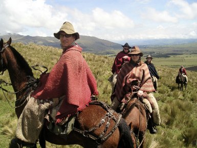 Reiter in traditioneller Kleidung genießen die Aussicht auf die grüne Landschaft am Cotopaxi, umgeben von Bergen und Wolken.