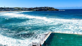 Der Pool am Bondi Beach reflektiert das Sonnenlicht, während Wellen an den Rand schlagen und Menschen schwimmen.