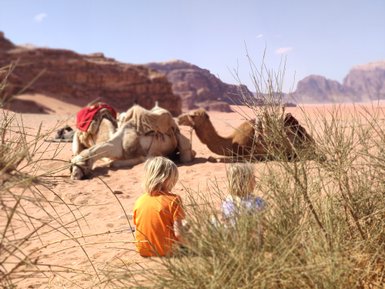 Zwei Kinder sitzen im Wadi Rum, während Kamele im Hintergrund auf dem Sand ruhen, umgeben von sanften Hügeln.