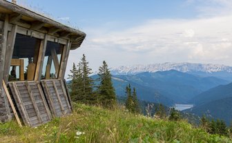 Ein rustikales Holzgebäude mit großen Fenstern steht auf einem Hügel, umgeben von grünen Wiesen und hohen Bergen im Hintergrund.