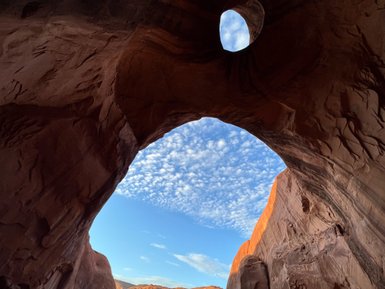 Ein beeindruckender Blick durch einen großen Felsenbogen auf den blauen Himmel mit Wolken und roten Felsen im Hintergrund.