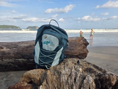 Am Strand von Playa Samara liegt ein Rucksack neben einem großen Holzstamm, während Kinder am Wasser spielen.