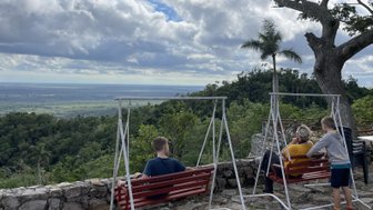 Drei Personen sitzen auf roten Schaukeln und genießen die Aussicht auf eine grüne Landschaft unter einem bewölkten Himmel.