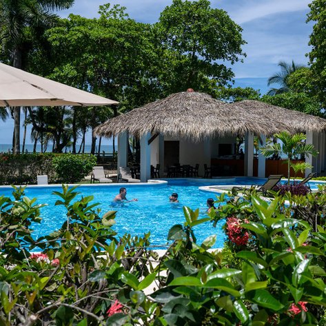 Ein einladender Poolbereich mit Palmen und einem Strohdach, wo Menschen entspannt im Wasser plaudern und lachen.