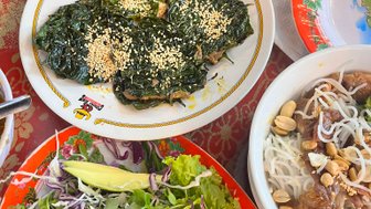 Ein Teller mit vier grünen, mit Sesam bestreuten Blättern, umgeben von frischem Salat und einer Schüssel mit Reisnudeln.