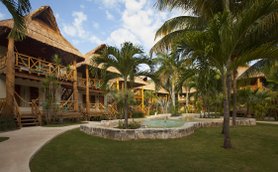 Ein tropischer Garten mit Palmen und einem kleinen Teich, umgeben von rustikalen Hütten mit Strohdächern.