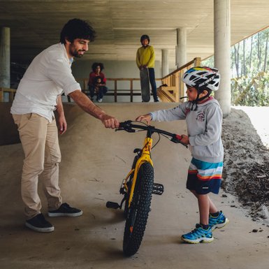 Ein junger Junge mit einem Fahrrad wird von einem Erwachsenen unterstützt, während andere Kinder im Hintergrund zuschauen.