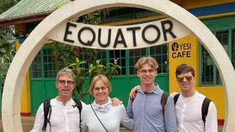 Eine Gruppe von vier Personen steht lächelnd am Äquator in Uganda, umgeben von bunten Gebäuden und Pflanzen.