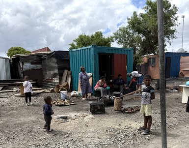 Eine Gruppe von Menschen steht vor bunten Containern in einem informellen Siedlungsgebiet, umgeben von Schutt und Steinen.