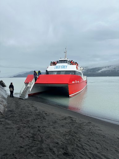 Ein rotes Boot mit der Aufschrift 'LAGO GREY' liegt am Ufer eines ruhigen Gewässers, während Passagiere an Bord gehen.
