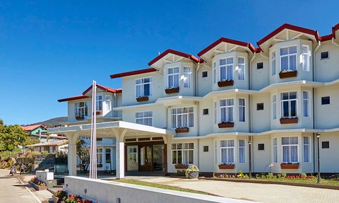 Ein modernes Hotelgebäude mit hellblauer Fassade und roten Dächern, umgeben von blühenden Blumenbeeten und klarem Himmel.