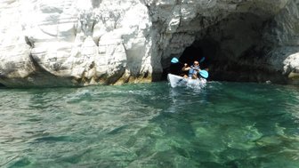 Ein Kajakfahrer paddelt durch klares Wasser in eine Höhle, umgeben von weißen Felsen und strahlendem Sonnenlicht.